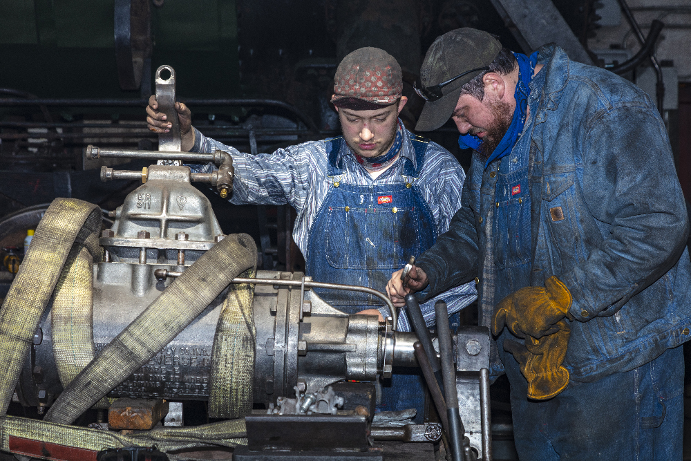 Two men in overalls working on a steam locomotive part.