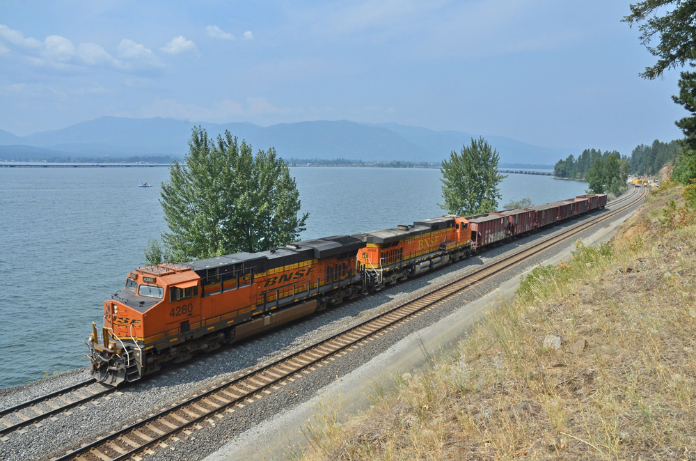Two orange locomotives with cars of ballast next to lake