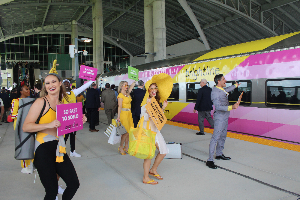 Costumed dancers welcoming a yellow and pink passenger train. Brightline