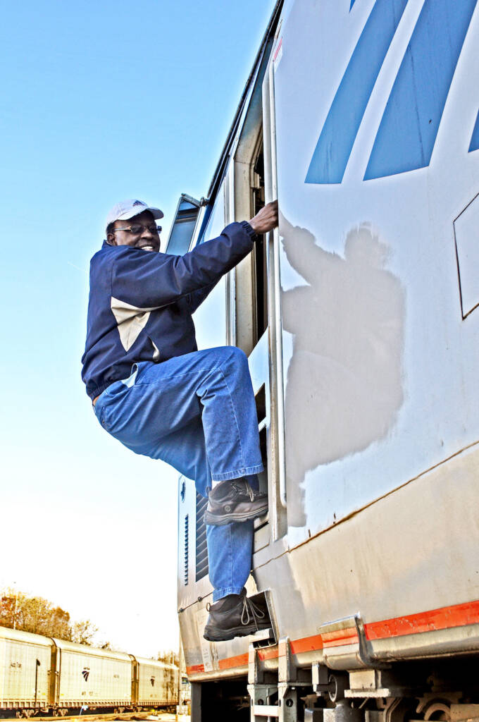 A railroad worker, dressed in blue jeans and a blue windbreaker, climbs the ladder to reach the cab of a passenger locomotive