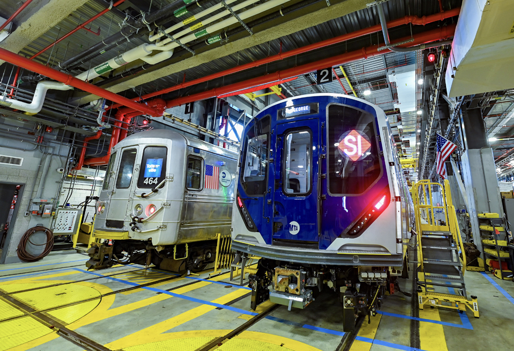 New subway car and old car sit on adjacent tracks in shop building