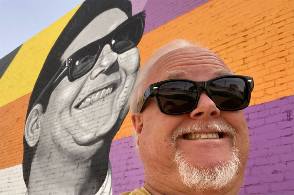 man smiling with glasses on in front of mural