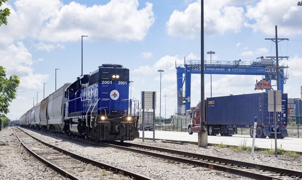 Train with blue diesel locomotive on track with gantry crane for port in background
