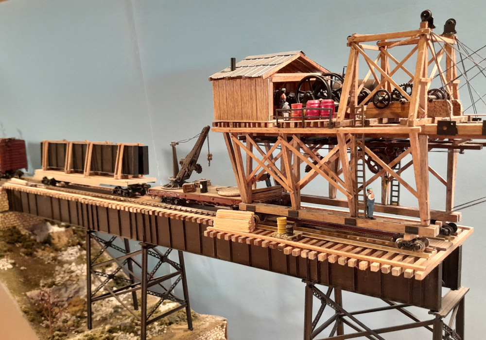 A multi-level wooden structure with a crane moves along a partially constructed wooden train trestle