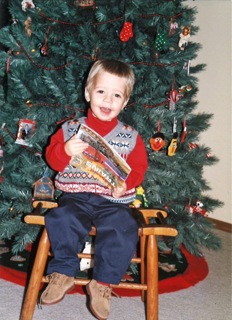 A little boy in front of a Christmas tree holding a book about trains upside down