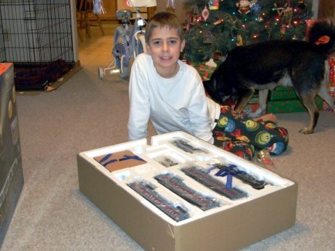 A young boy on the floor beneath a Christmas tree unboxing a train set