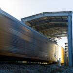 A train passes through a tall, gray, open-ended shed