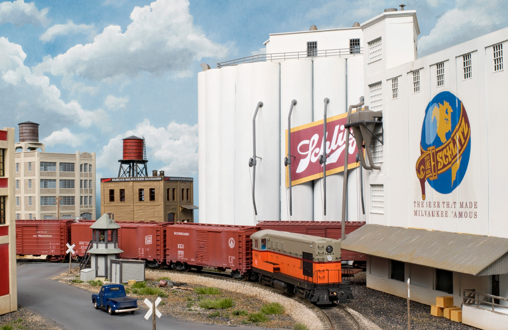 An orange-and-gray end-cab diesel leads red boxcars past towering white grain elevators bearing Schlitz logos
