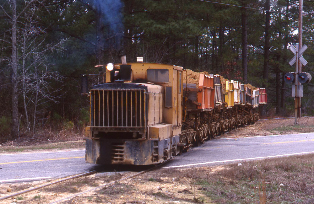 A small yellow industrial locomotive pulls a short train of multi-colored side-dump hoppers across a road crossing