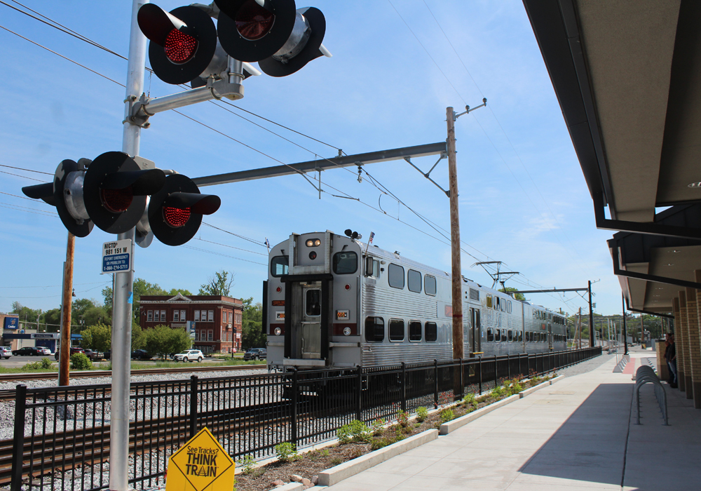 Two-car bilevel train arrives at station, with crossing signals in foreground