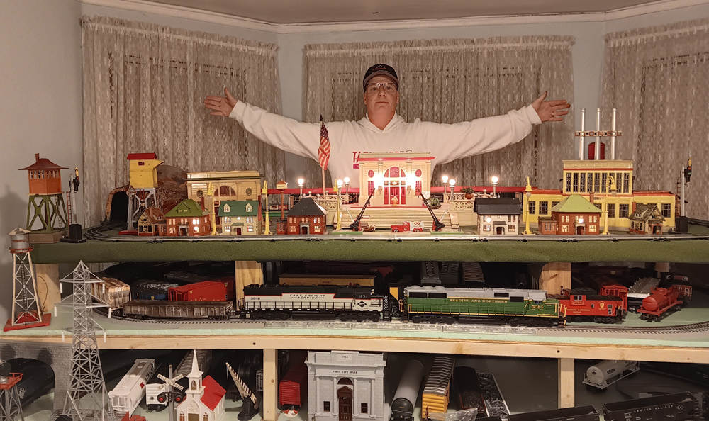 man behind toy train layout, stretching his arms out wide