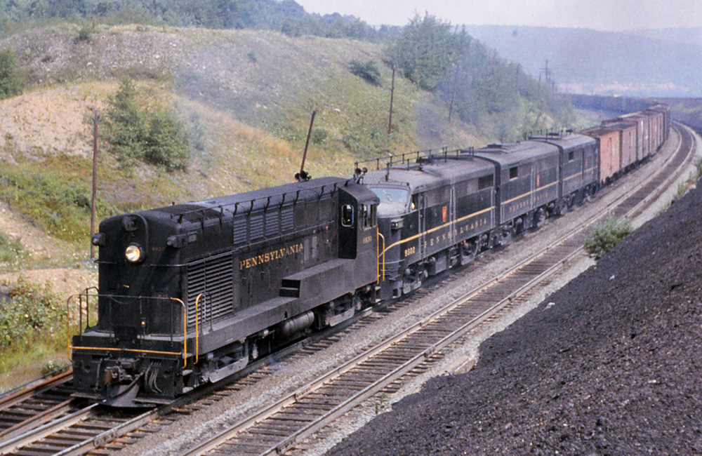 Four dark green diesel locomotives with Pennsylvania RR Trainphone antennas on top round a curve with a train of assorted freight cars.