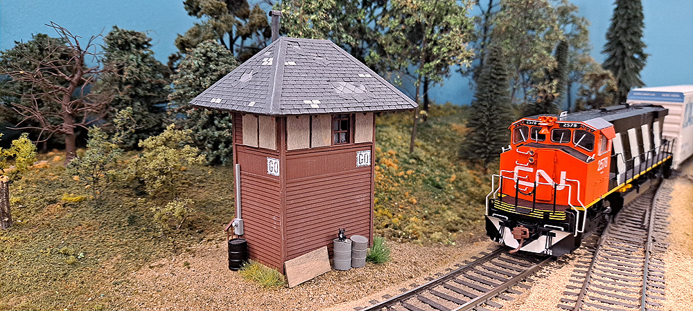An HO scale model of a railroad interlocking tower with boarded-up windows stands next to a track as a red-nosed Canadian National wide-nose Alco M420 approaches from the right