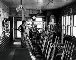 Black and white photo of the inside of an interlocking tower showing floor-mounted levers with a young man in T-shirt and jeans standing at the end of the row of levers.