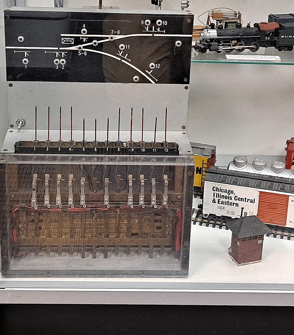 Model interlocking machine with black model board showing track layout next to HO scale model of a red interlocking tower.