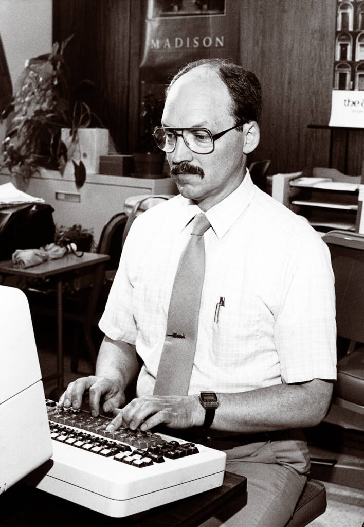 Black-and-white photo of man wearing glasses, dress shirt, and necktie typing at a word processor. 
