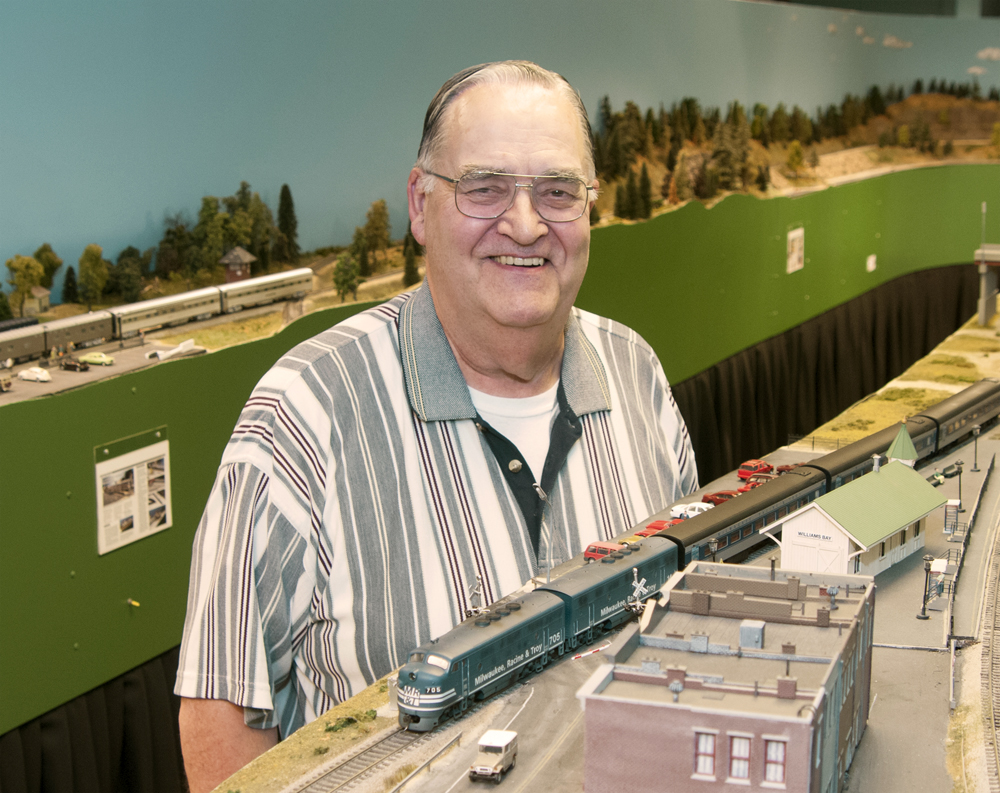Color photo of man wearing glasses in striped polo shirt standing by model railroad.