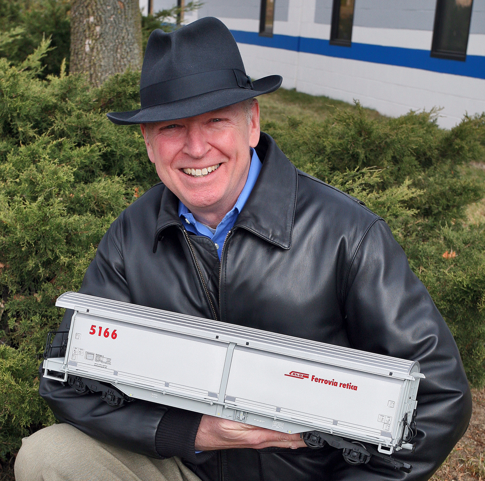 Color photo of man in leather jacket with dress hat holding a large scale train.