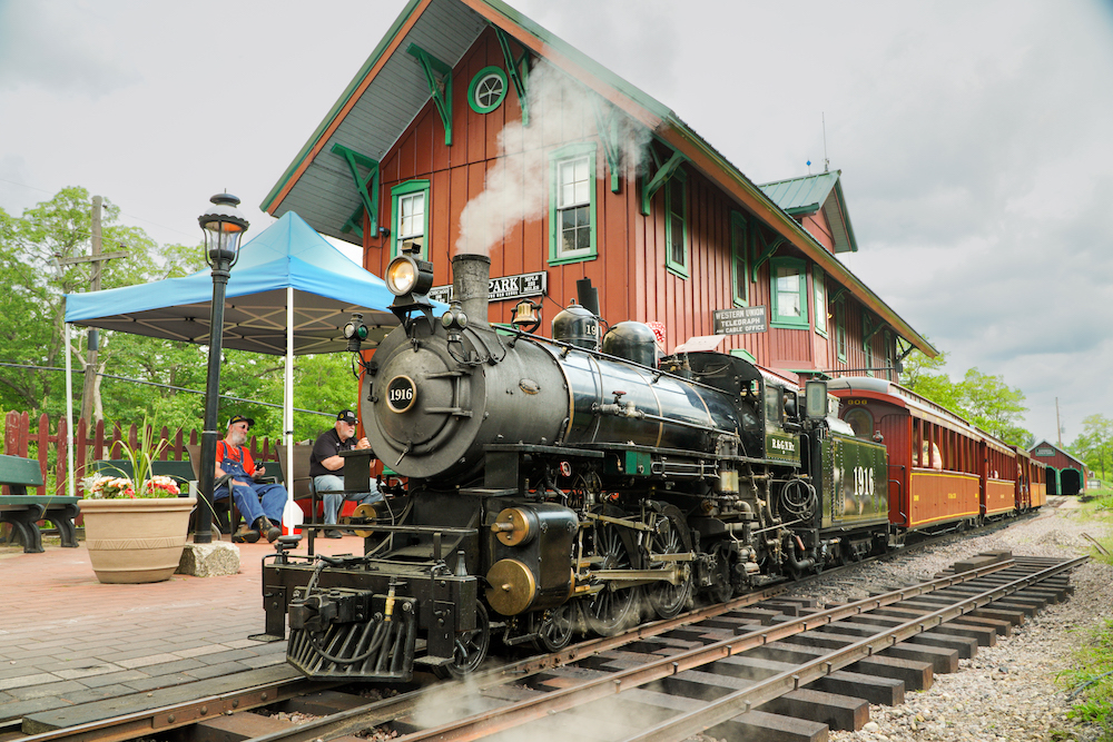 Little steam-powered train sits at two-story train station on a cloudy day.