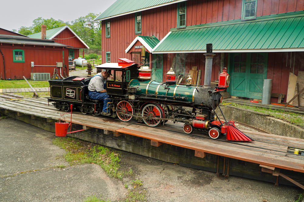 Little 4-4-0 steam locomotive sits in turntable with red-painted buildings in the background.