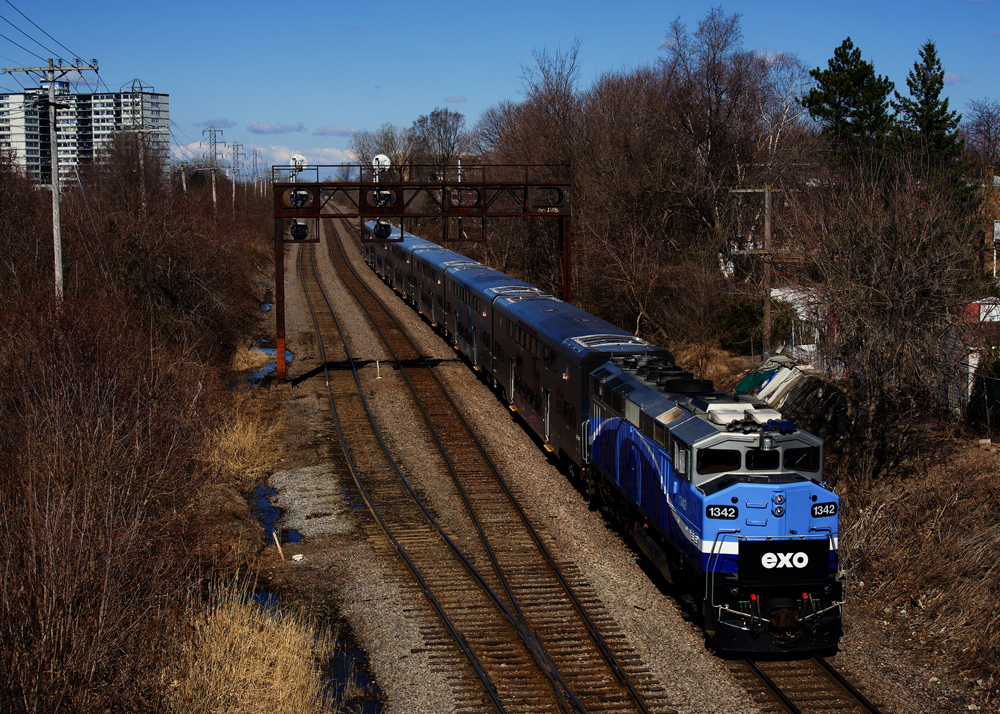 Blue and silver locomotive with white trim pulling six cars of bilevel passenger equipment past signal bridge