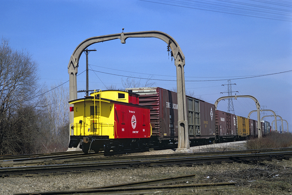 Color photo of red-and-yellow caboose and boxcars going under concrete arches.