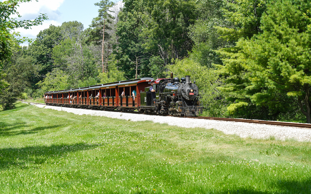 Little steam locomotive pulls a long string of wooden passenger cars.