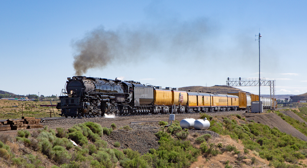 Steam locomotive with passenger and freight cars passes under signal bridge on high plains.