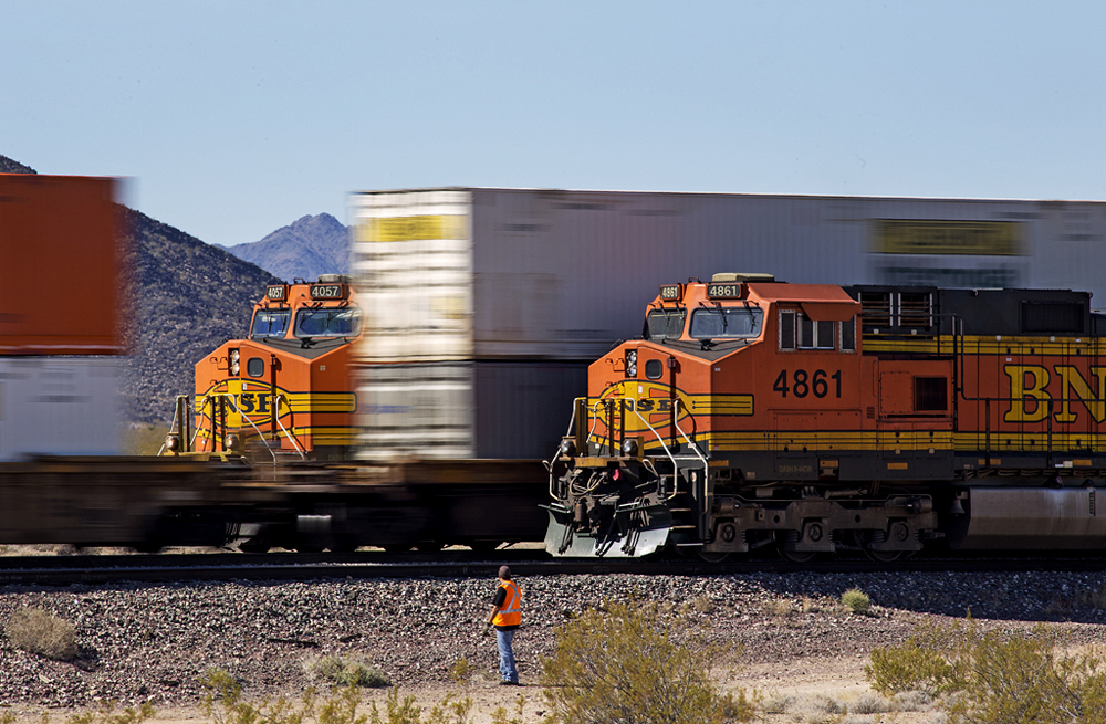 Crew member stands by tracks as trains pass in desert landscape.