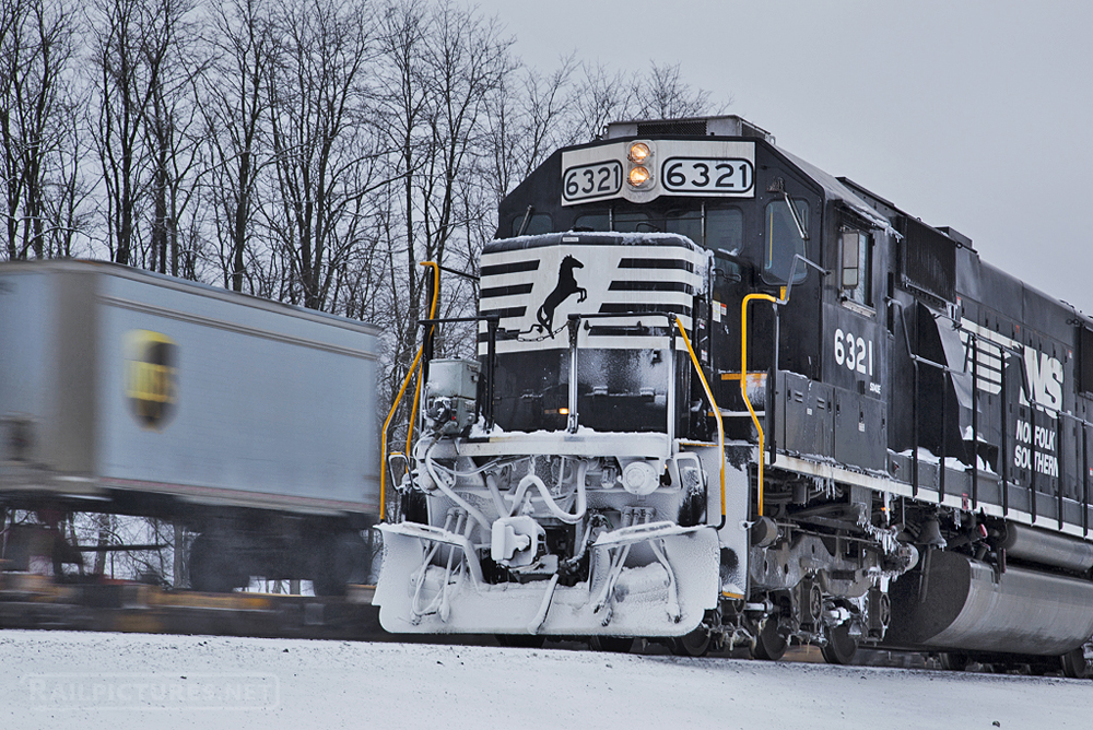 Black and white locomotive in snow with piggyback cars passing on adjacent track