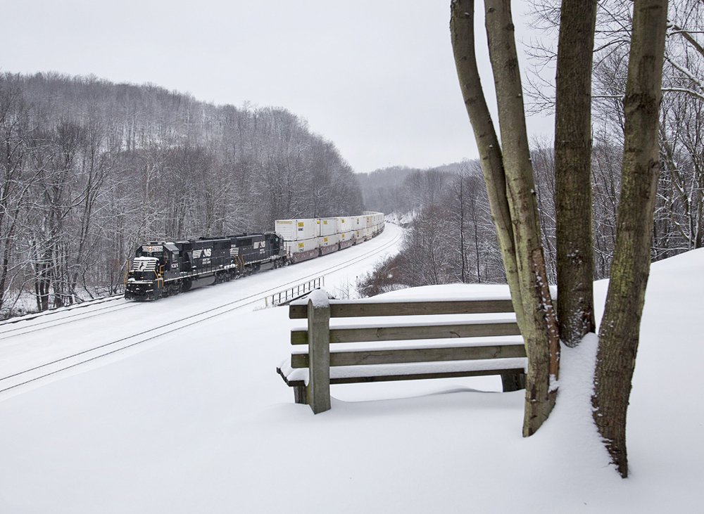 Intermodal train with black locomotives passes bench covered with snow