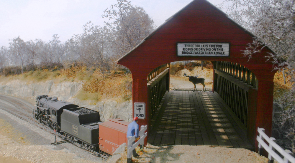 A red covered bridge on a model railroad layout with a man on one end and a moose on the other with a train passing under the bridge