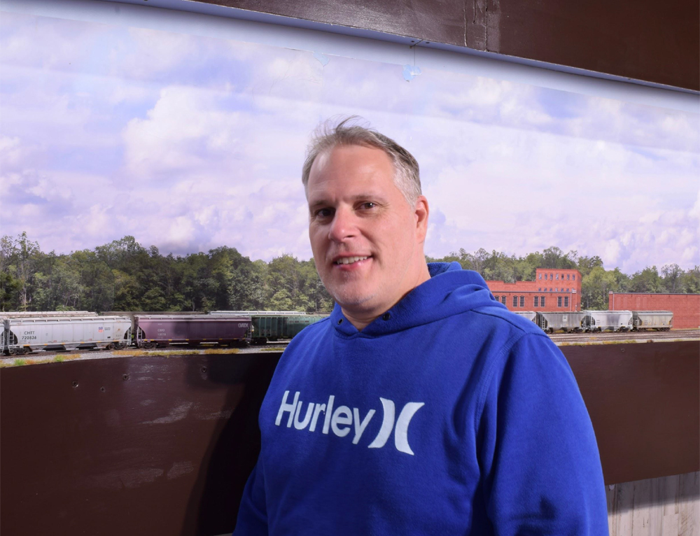 Man in blue sweatshirt standing next to model train layout