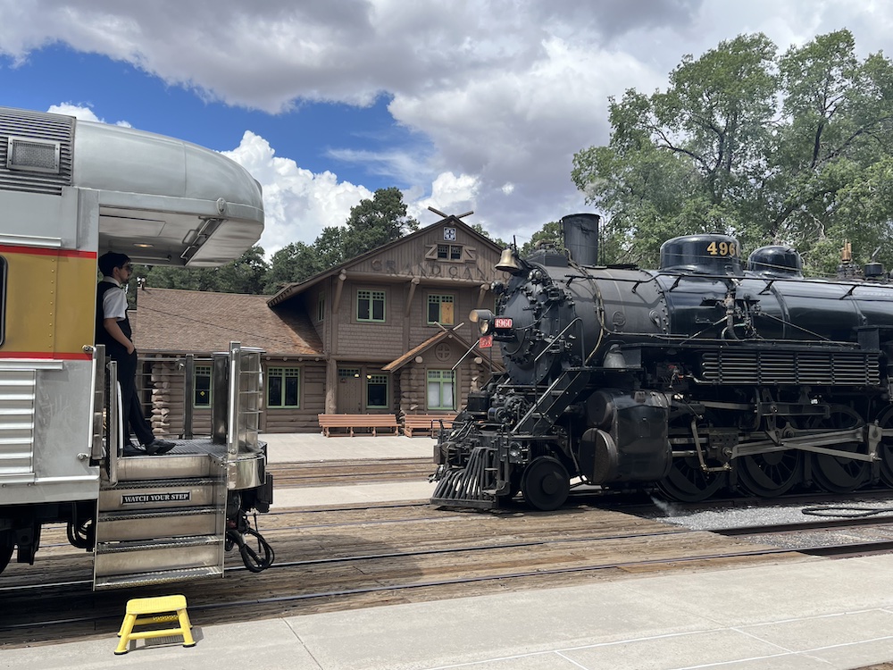 Steam locomotive sits in front of log-cabin depot