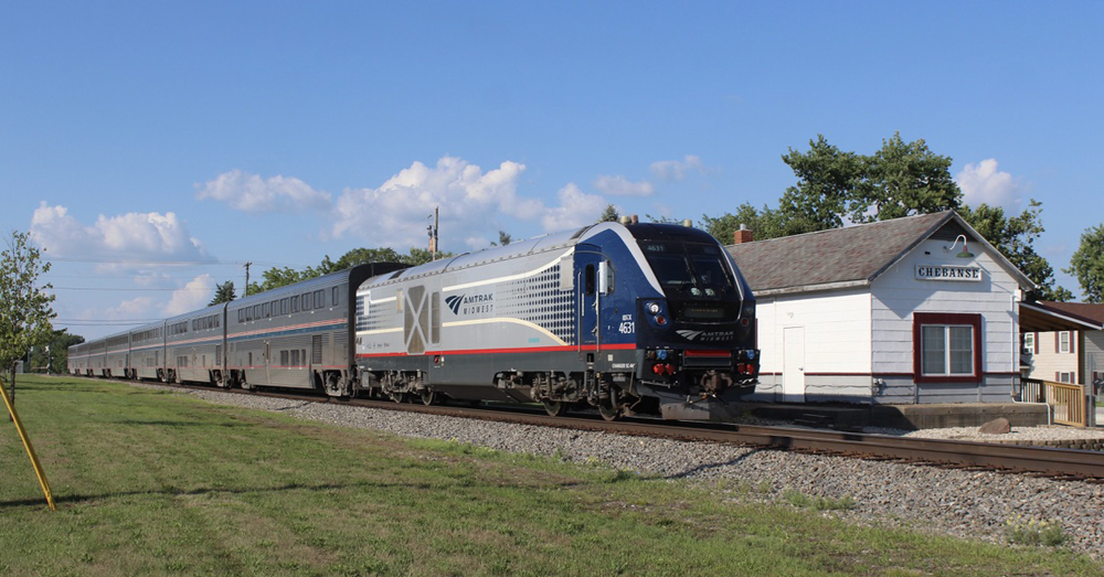 Train with Amtrak Midwest locomotive and Superliner cars passing small-town station