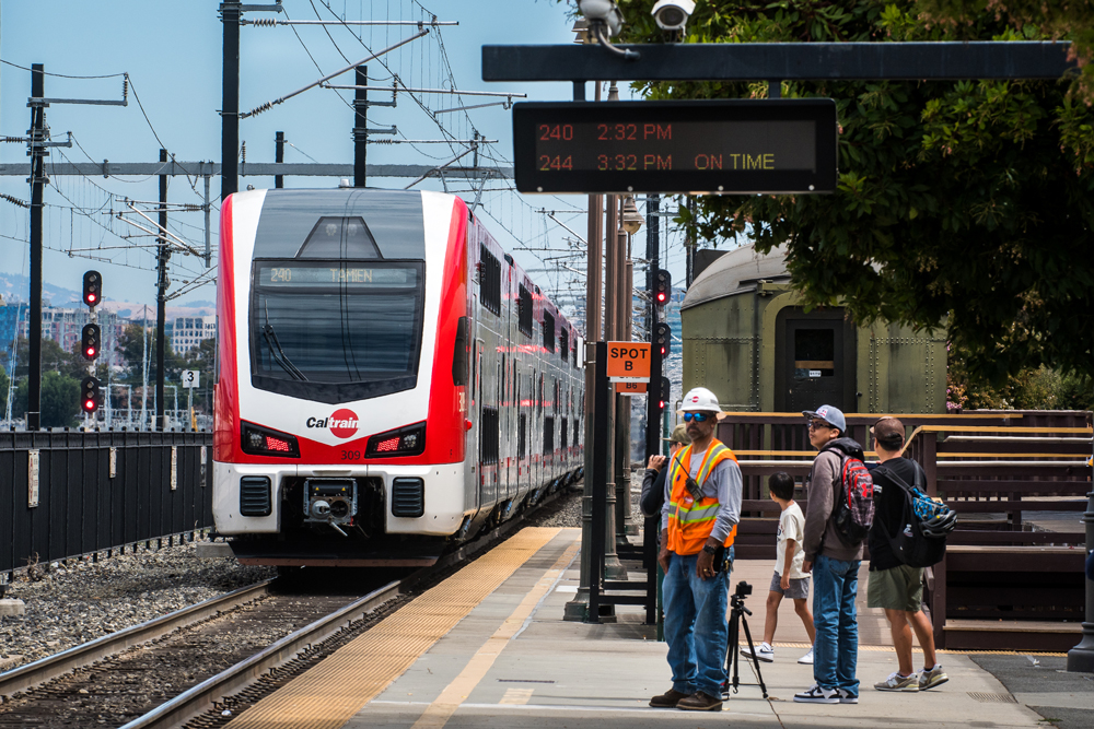 White and red electric train arrives at station platform where several people are watching