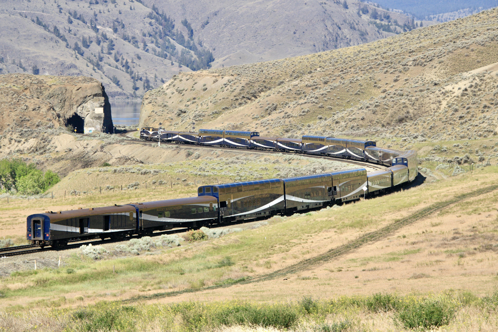 Long passenger train of dark blue single- and billevel cars with gold and white trim in rolling hills.