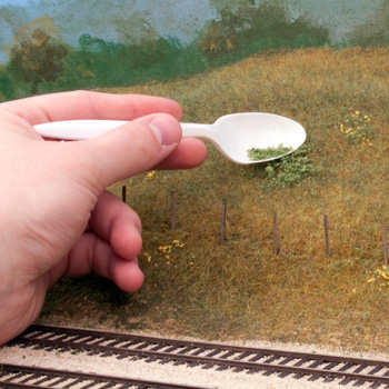 A hand holding a spoon sprinkles ground foam on a model landscape