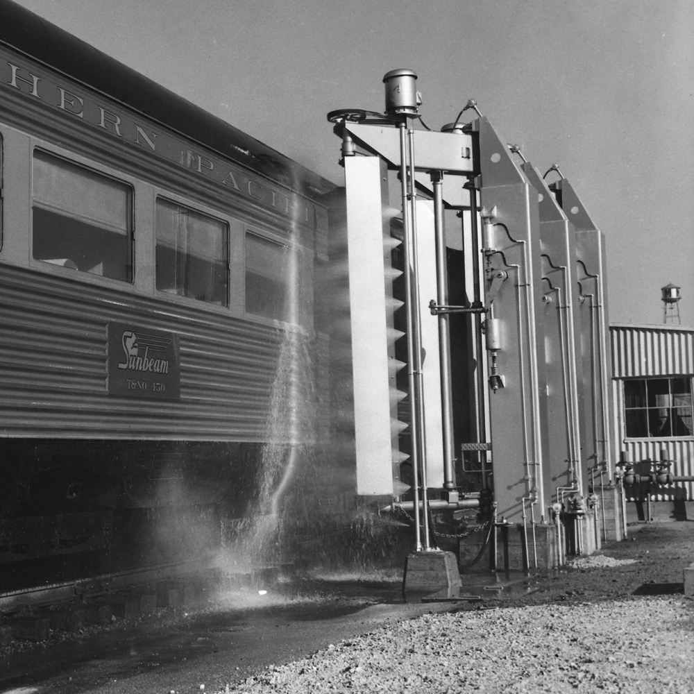 Black-and-white image of streamlined passenger car by trackside mechanical washer.