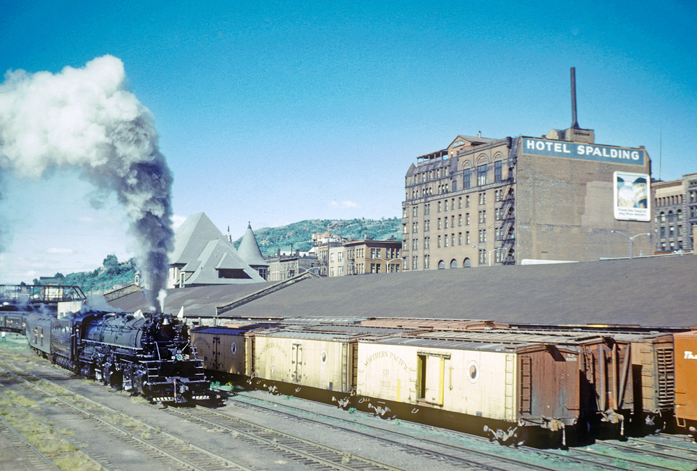 Large steam locomotive passes yard track of freight cars