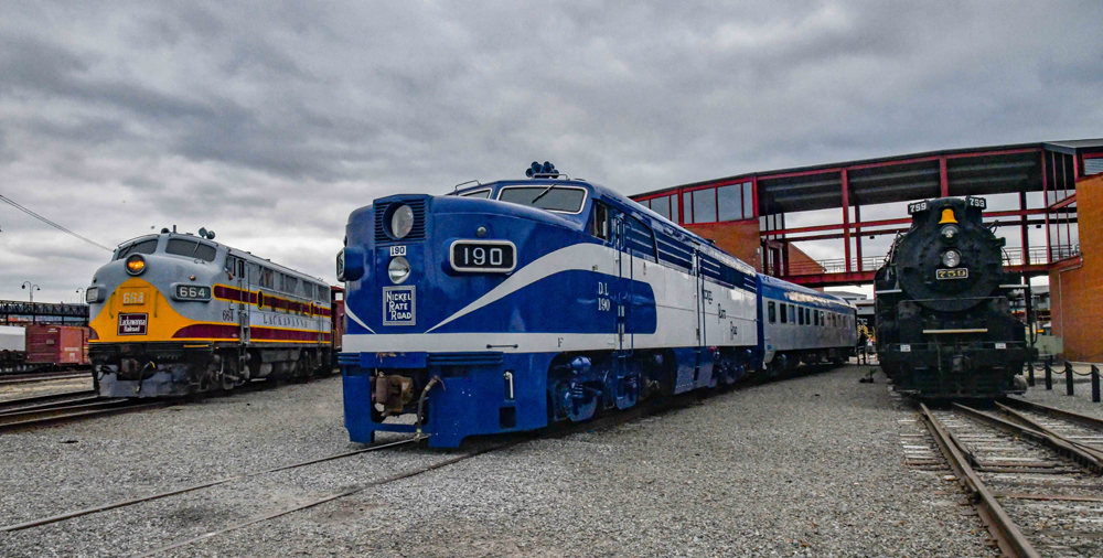 Two diesel cab units and steam locomotive on display under cloudy skies