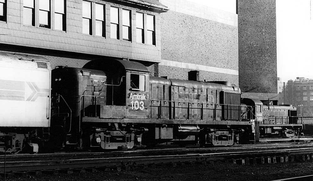 A black and white photograph of a locomotive in front of a brick building in a train yard