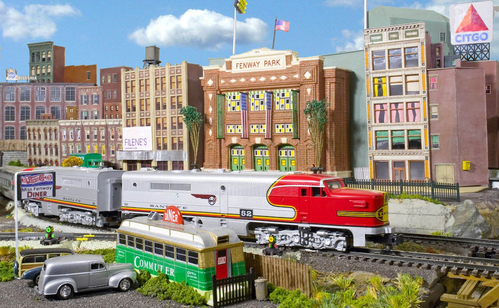 A silver and red toy train locomotive in front of a city skyline on a toy train layout