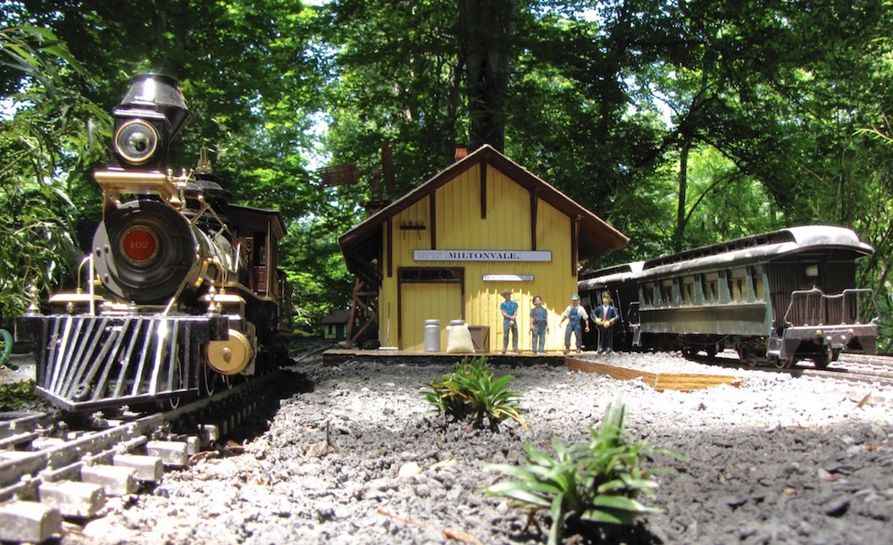 low angle photo showing steam locomotive, depot, and passenger car on garden railway