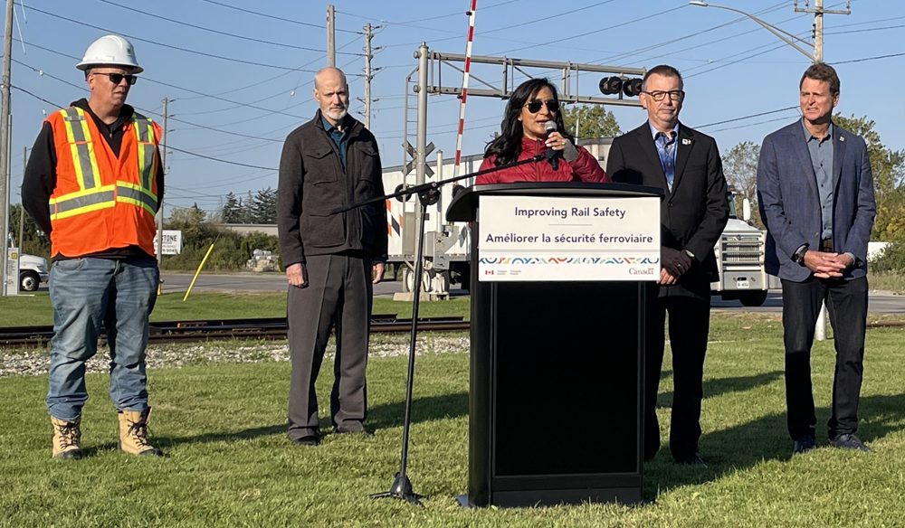 Woman speaking at podium near grade crossing as four men look on