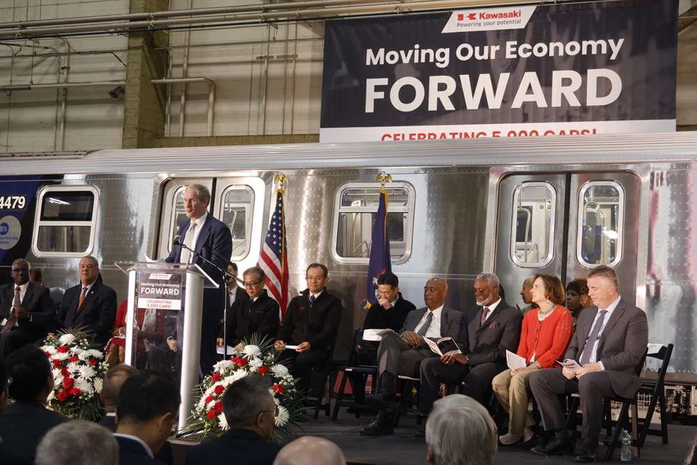 Man at podium with officials seated in background in front of subway car