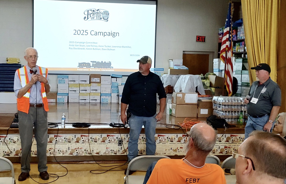 Man in orange vest speaking in front of projector screen