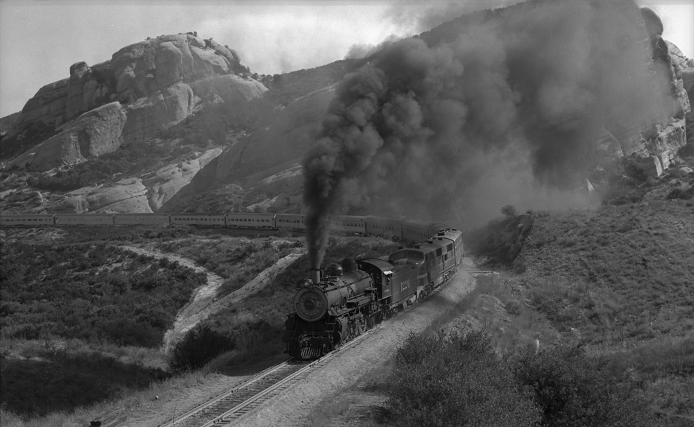 Black-and-white image of steam engine assisting streamlined passenger train in Southern California mountains