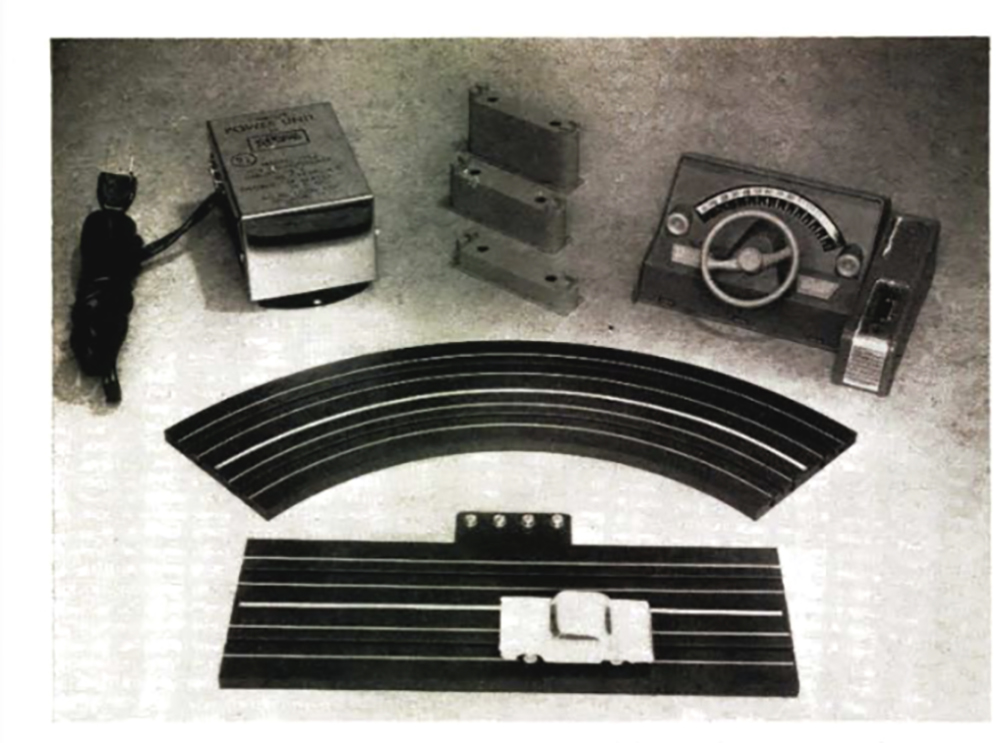 Black and white photo showing a straight and curved section of black model roadway with white center stripes, a small white car, a gray control panel with small tan steering wheel, and a metal power supply cube with black cord wound up next to it. 