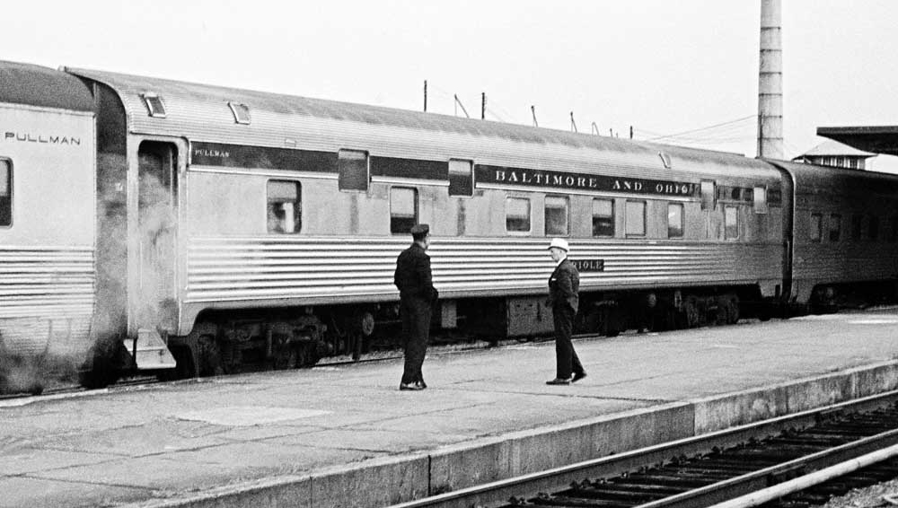 A black and white photograph depicting two men standing on a passenger platform in front of a budd slumbercoach passenger car